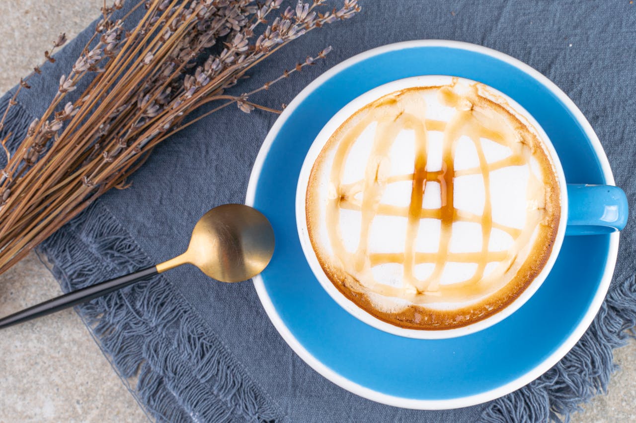 gallery-01 Artistic top view of a caramel-topped coffee in a blue cup, styled with dried flowers and gold spoon. Perfect for cozy moments.