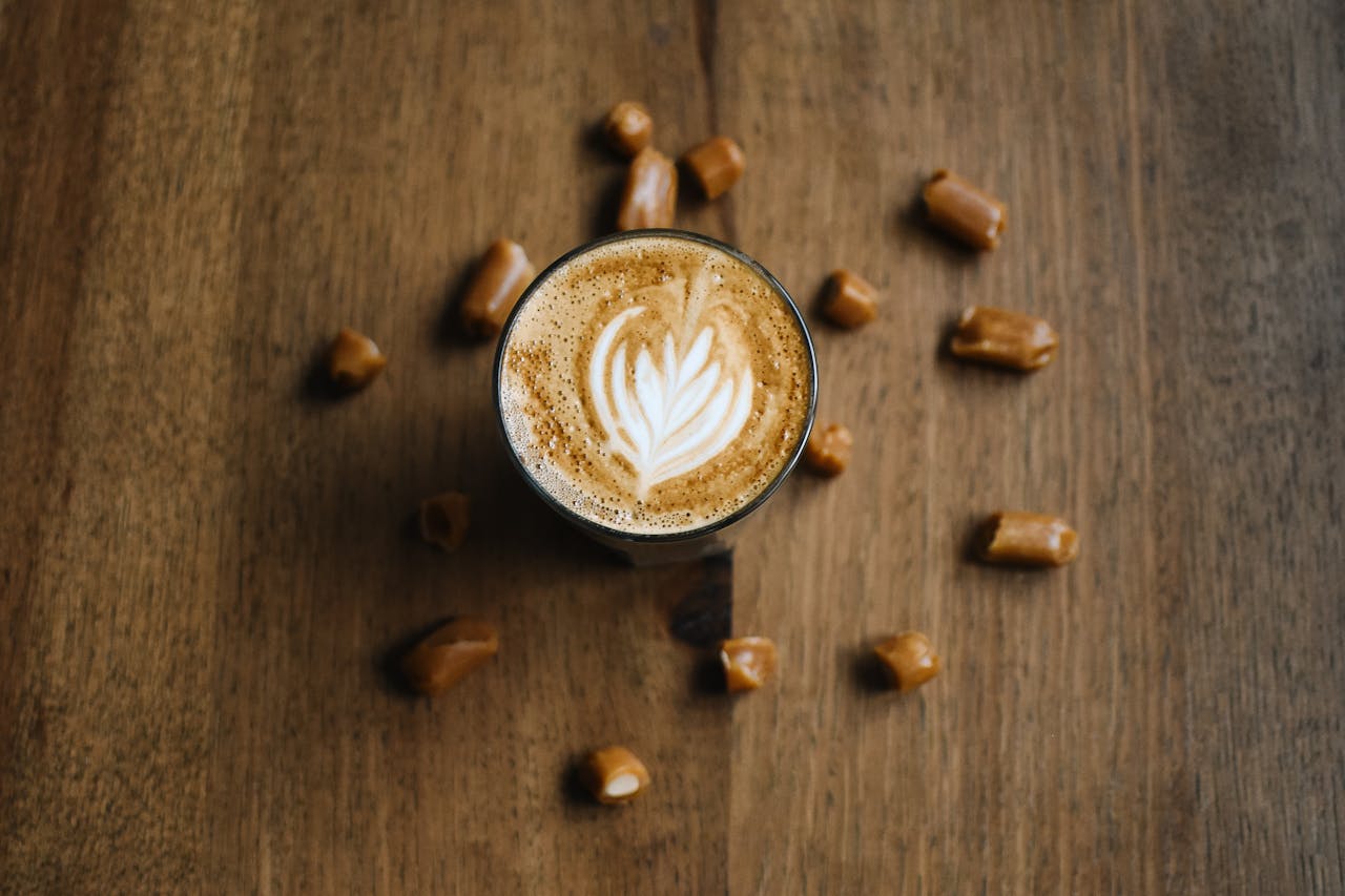 home-img A latte with intricate froth art surrounded by caramel candies, shot from above on a rustic wooden table.